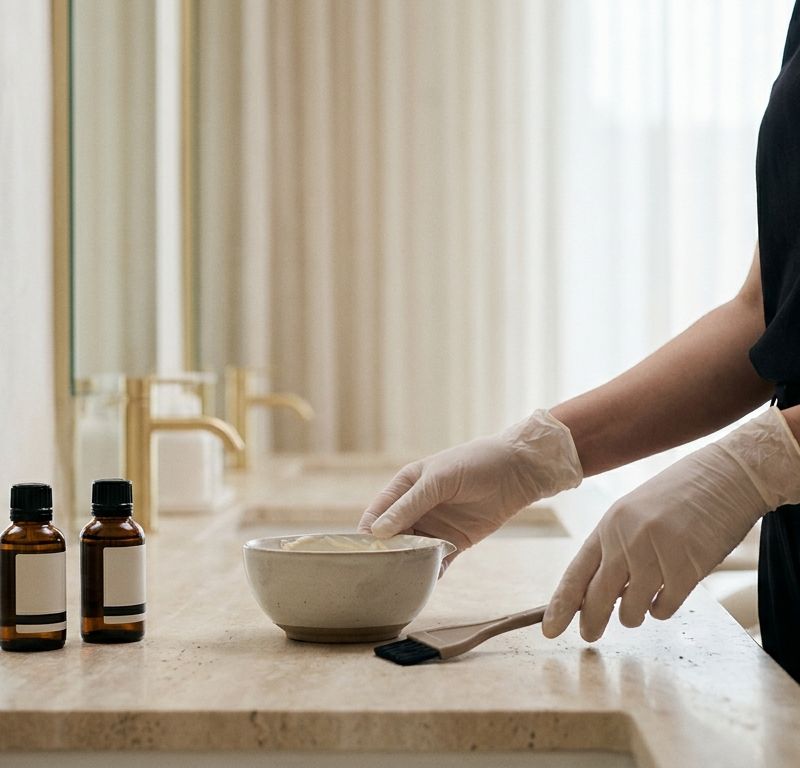 Color professional at travertine counter with bowl and brush, editorial warm salon, formula calculated before mixing