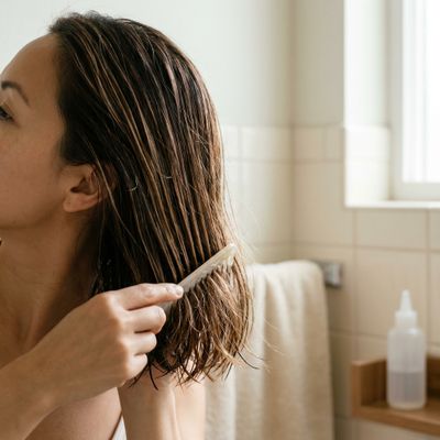 Una persona de perfil se pasa un peine de púas anchas por la melena castaña mojada, en un baño con luz de ventana y un bote sin marca en la repisa