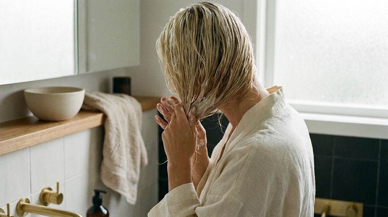 A woman in her own bathroom, seen from behind, working hair mask through the mid lengths and ends of her wet blonde hair
