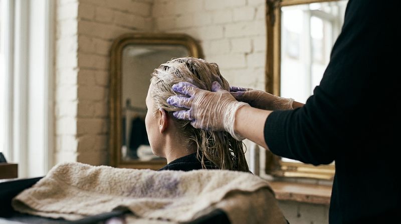 Uma mulher no banheiro de casa, de costas, distribuindo espuma violeta pelo próprio cabelo loiro molhado com luvas