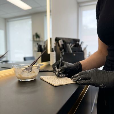 Colorista profissional revisando a ficha do cliente ao lado do bowl de fórmula antes de aplicar coloração para cobertura de fios brancos