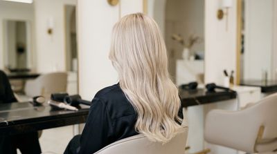 Cabelo champagne blonde dimensional, loiro pastel neutralizado com reflexos prateados finos, fotografado de costas em salão premium com luz natural
