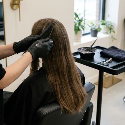 Manos con guantes negros abriendo una raya con un peine de cola sobre el cabello largo, castaño natural y sin color previo, de una persona sentada de espaldas en un salón, con un bol de crema ceniza al fondo