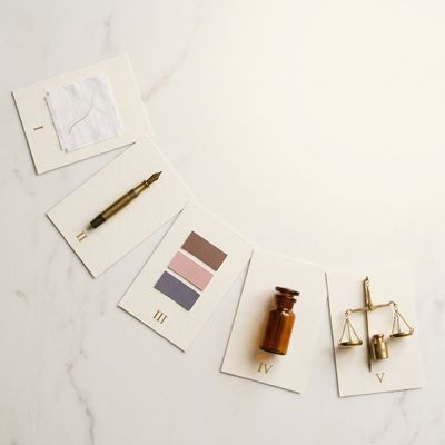 Flat-lay editorial em mármore creme com cinco objetos numerados: mecha, pena, paletas de cor, frasco de química, balança de precisão