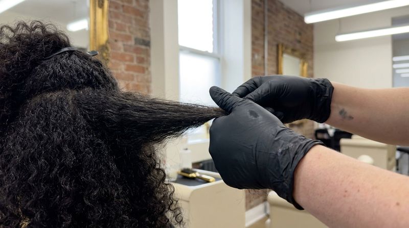 Gloved hands parting a section of dry curly hair backlit to assess uneven shine before bleaching