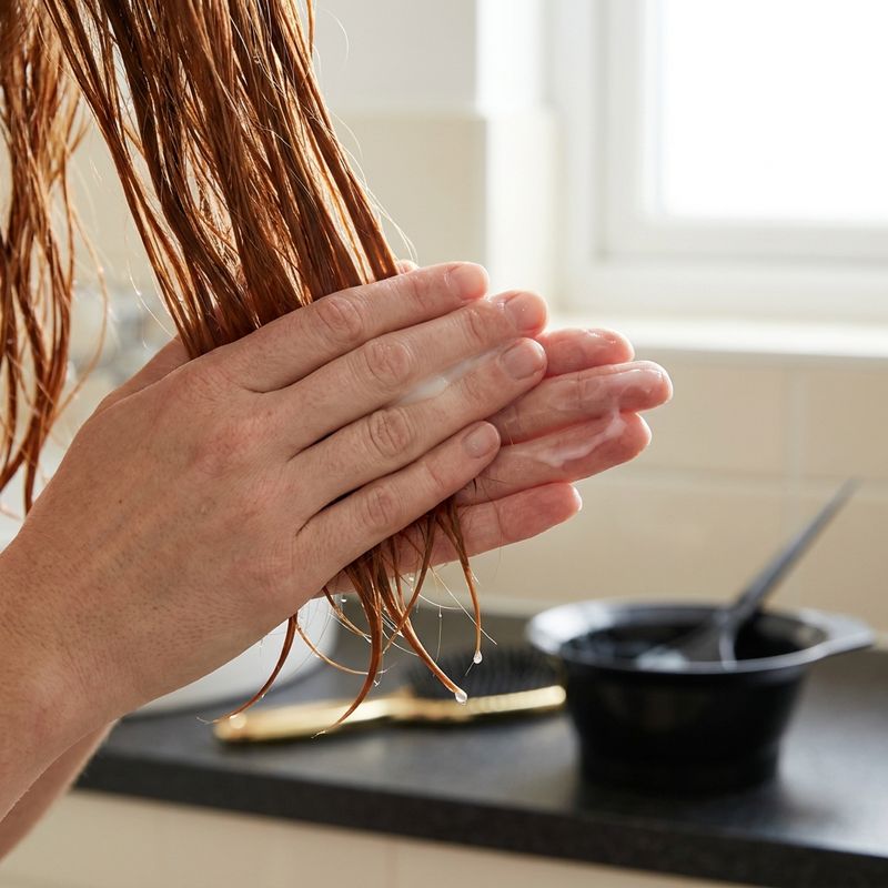Mãos aplicando condicionador do meio às pontas de um cabelo colorido acobreado, com um bowl de mistura desfocado ao fundo