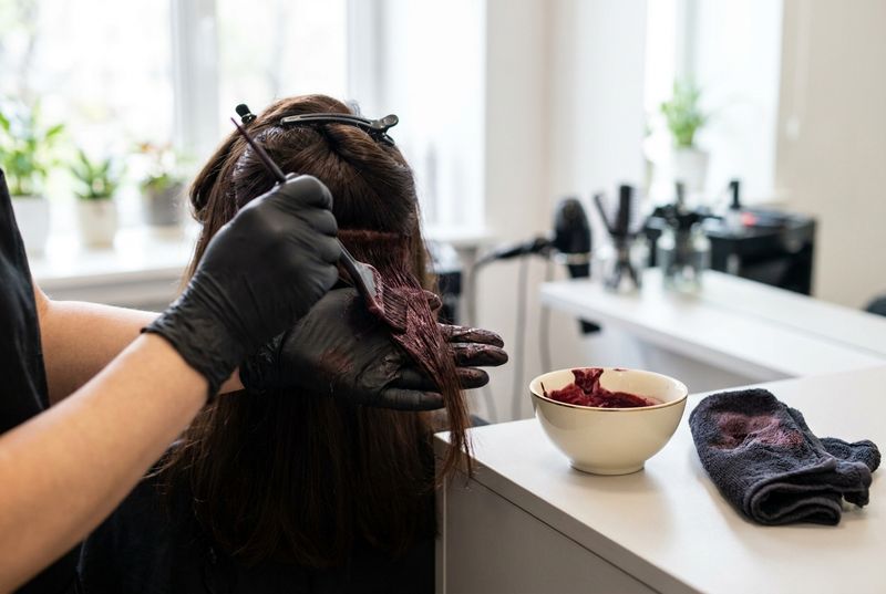 Hands in black gloves applying a dark violet-red color cream to a section of hair with a tint brush