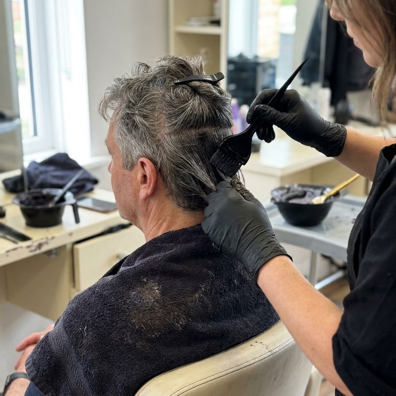 Gloved hands applying dye with a tint brush to salt-and-pepper grey hair at a client's nape, in a real salon with a mixing bowl in the background