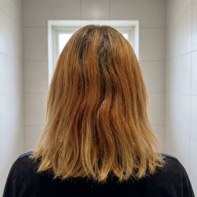 Cabelo com tons laranja desiguais depois de uma descoloração em casa, visto de costas no banheiro