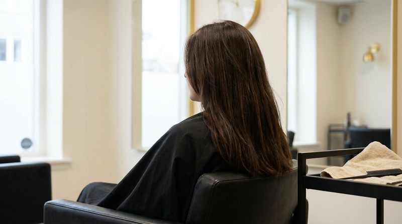 Persona de espaldas y casi de perfil, sentada en un sillón negro junto a una ventana, con una melena castaño chocolate profundo y uniforme