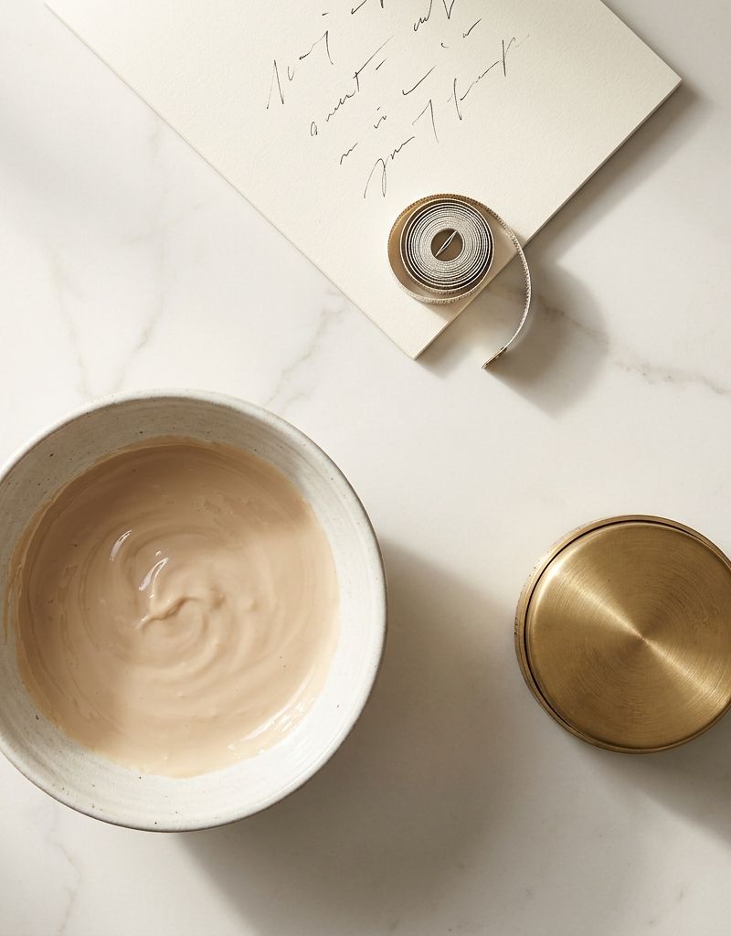 Bowl de colorante mezclado junto a temporizador analógico dorado y ficha técnica en superficie de mármol crema