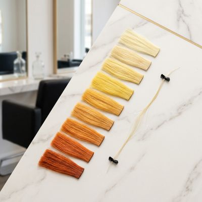 Colorista profissional avaliando uma mecha úmida com teste de elasticidade, ao lado de mechas de diferentes níveis de base de clareamento