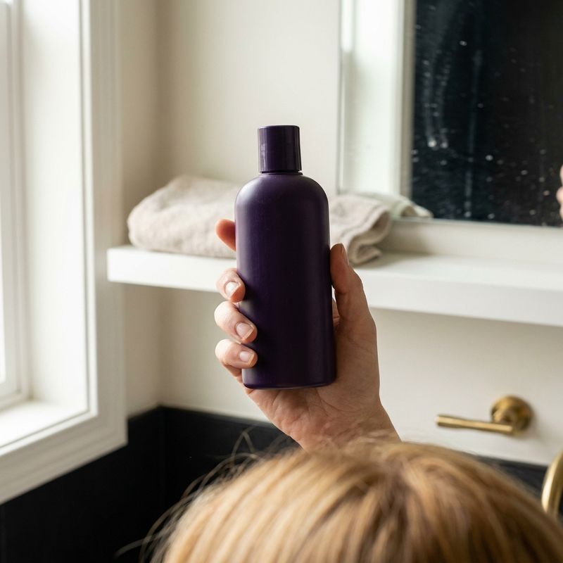Mano sosteniendo un bote de champú morado sin marca en un baño con toalla usada y luz de ventana