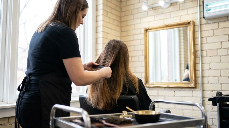 Colorist combing chestnut brown hair forward under salon light to compare the tone at the roots, mid-lengths and ends