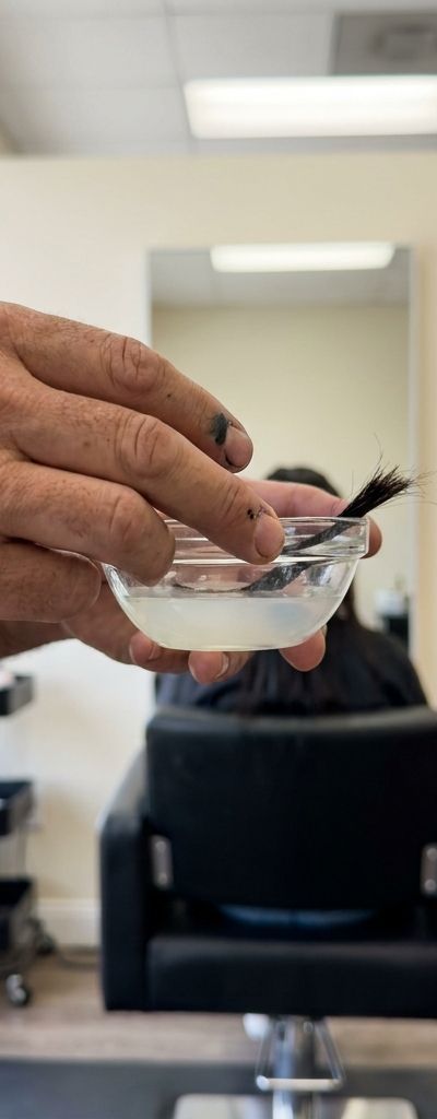 Manos de un profesional del color sosteniendo un bol de cristal con un mechón sumergido, con una persona esperando al fondo del salón