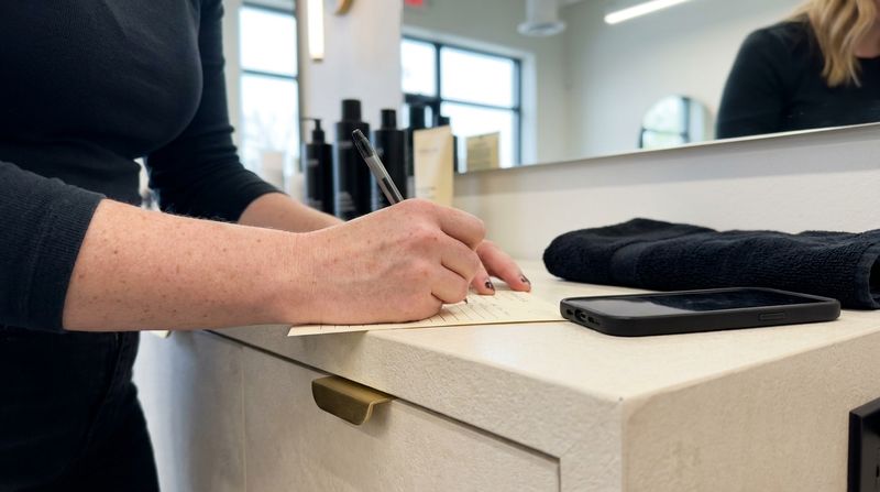 Manos anotando unas líneas en una ficha de cliente sobre el mostrador del salón, con un móvil al lado