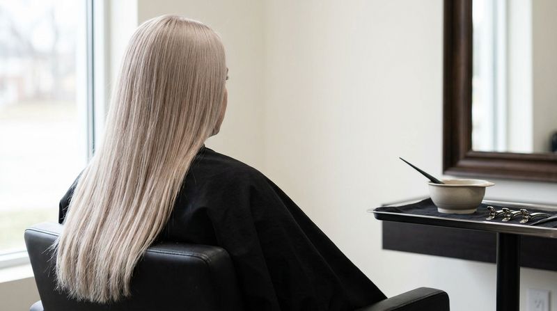 An even cool beige blonde from roots to ends, seen from behind in a salon chair, with a black cape and natural window light