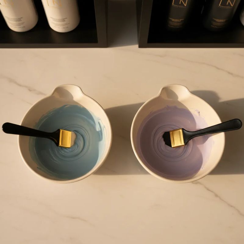 Duas tigelas de mistura comparando toner com pigmento azul (esquerda) e toner com pigmento violeta (direita) sobre superfície de mármore creme